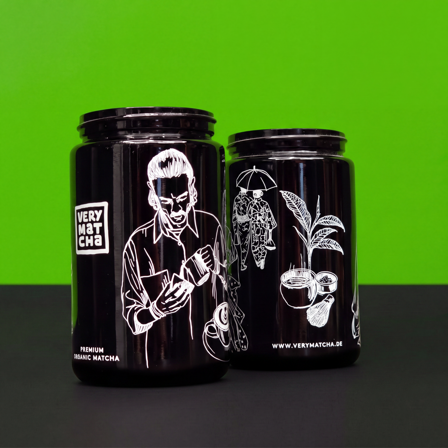 Zwei schwarze Matcha-Gläser mit weißem Siebdruck vor grünem Hintergrund